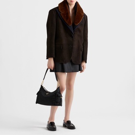 Model carrying Prada Accessories black shoulder bag with top handle, styled with dark brown faux fur collar blazer and mini skirt.