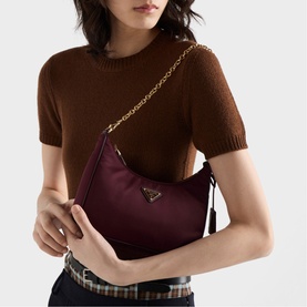 Model carrying Prada Accessories burgundy nylon hobo bag with gold chain strap and triangle logo, styled with brown knit top.
