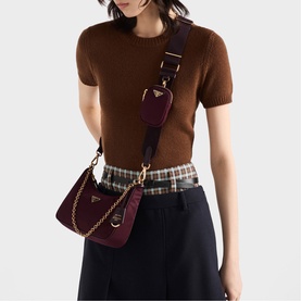Model carrying Prada Accessories burgundy crossbody bag with gold chain strap and logo detail, styled with brown knit top and plaid skirt.
