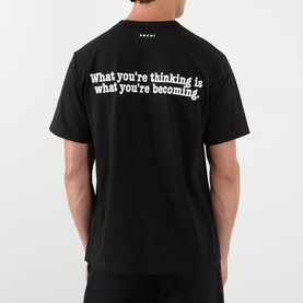 Model wearing Sacai black cotton T-shirt, back view, with white slogan print reading "What you're thinking is what you're becoming.