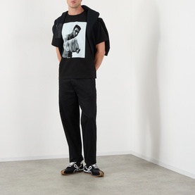 Model wearing SACAI black cotton T-shirt with monochrome graphic print, styled with black trousers and dark trainers.