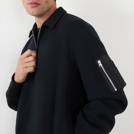 Model wearing SACAI black polo with half-zip collar and silver-zip nylon sleeve pocket detail, close-up shot.