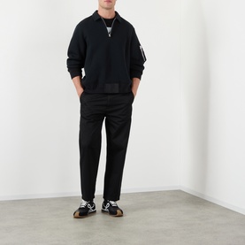 Model wearing Sacai black cotton-blend polo with zip collar and nylon sleeve panel, styled with black trousers and monochrome trainers