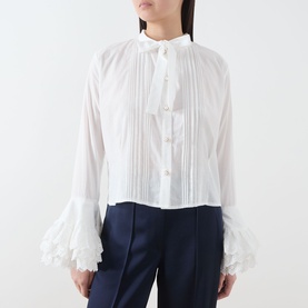 Model wearing Sister Jane white cotton blouse with pintuck front, ruffle cuffs and neck tie, styled with navy wide-leg trousers.