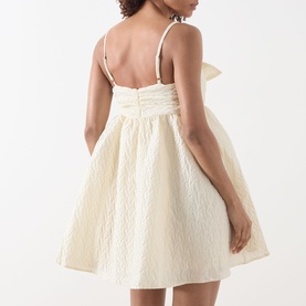 Model wearing Sister Jane Honey Jacquard Mini Dress in cream, showing back bow detail, spaghetti straps, and flared jacquard skirt.
