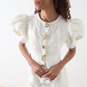 Model wearing Sister Jane Milos Brooches Mini Dress in white jacquard with puff sleeves and gold embellished brooch buttons at front.
