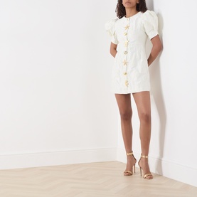 Model wearing Sister Jane Milos Brooches Mini Dress in white jacquard with puff sleeves and gold embellished buttons, styled with gold heeled sandals