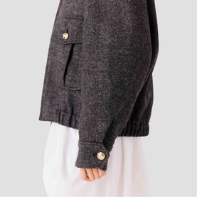Model wearing Maje dark grey textured jacket, close-up of sleeve with gold button cuff detail and chest pocket, styled over white dress.