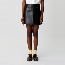 Model wearing Claude Pierlot black faux leather mini skirt with gold-tone button detail, styled with white shirt, yellow socks and dark brown loafers.