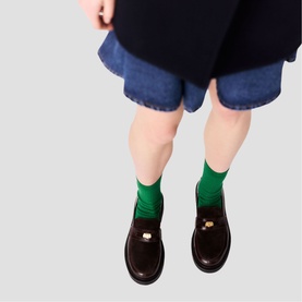Model wearing Maje dark brown loafers with gold-tone hardware, styled with green socks and navy blue shorts