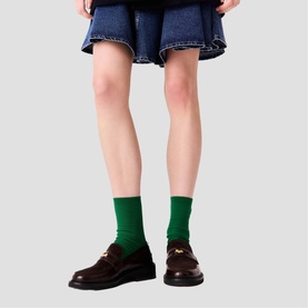 Model wearing Maje dark brown leather loafers with gold-tone hardware, styled with green crew socks and a denim mini skirt.