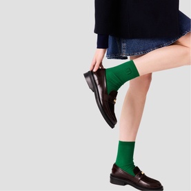 Model wearing Maje dark brown leather loafers with gold-tone hardware, styled with green ankle socks and a navy mini skirt.