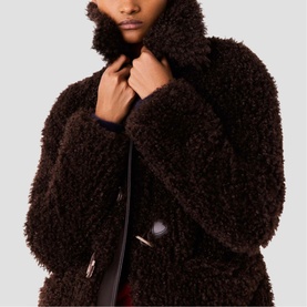 Model wearing Maje dark brown teddy fleece coat, hands raised to collar, toggle fastenings visible at front