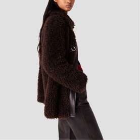 Model wearing Maje dark brown curly faux fur jacket, styled with red top and black leather trousers, side view