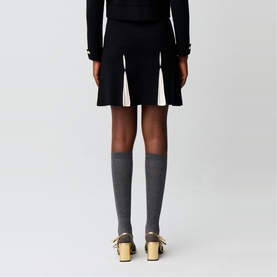 Model wearing Claude Pierlot black pleated mini skirt, styled with black knit top, grey knee-high socks and gold mule heels, rear view.