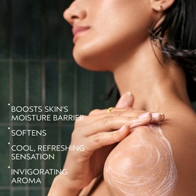 Model applying Aveda Rosemary Mint Invigorating Body Wash to shoulder, with lather visible against wet skin in a shower setting.