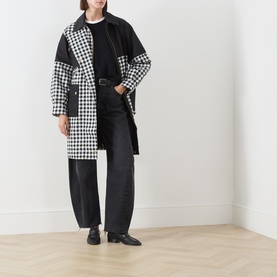 Model wearing Barbour International Gea Gingham Showerproof Coat in black, longline with patchwork gingham print, belted, styled with dark wide-leg