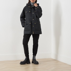 Model wearing Barbour International Molina Showerproof Coat in black with gold snap buttons, detachable hood, styled with black skinny trousers and