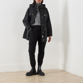 Model wearing Barbour International Molina Showerproof Coat in black with detachable hood and gold snap buttons, styled with black cargo trousers and