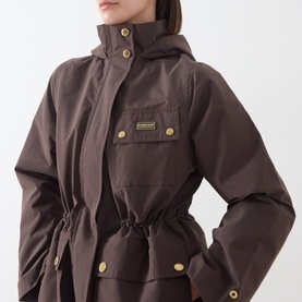 Model wearing Barbour International truffle coat with drawstring waist, chest pocket, gold-tone buttons and snap fastenings, and hood detail.