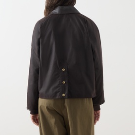 Model wearing Barbour International dark brown waxed cotton jacket, back view, gold snap buttons, leather collar detail, styled with olive trousers