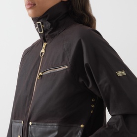 Model wearing Barbour International dark brown cotton jacket with gold-tone zip and hardware detail, high collar with buckle strap visible.