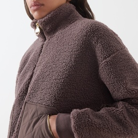 Model wearing Barbour International truffle teddy fleece jacket with high funnel neck, gold zip, and quilted hem trim detail.