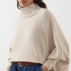 Model wearing Barbour International moonbeam roll-neck knit sweater with batwing sleeves and gold buttons, styled with blue jeans and tan belt.