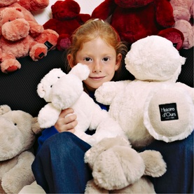 Child surrounded by assorted plush teddy bears, holding Dou Dou beige plush bear with branded label, alongside pink and red soft toys