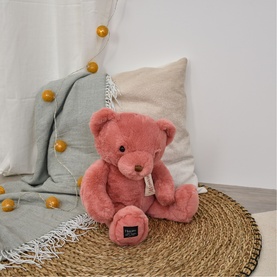 Dou Dou pink plush teddy bear, seated on a wicker tray with linen cushions and gold ball fairy lights in background.