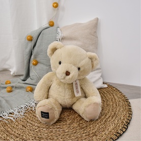 Dou Dou cream plush teddy bear, seated on a woven mat with cushions and a knitted throw in the background