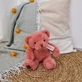 Dou Dou pink plush teddy bear with label tag, seated against neutral cushions on woven surface