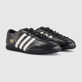 Adidas low-top trainers in core black with cream white three-stripe detail, perforated leather upper, black rubber sole, gold branding script.