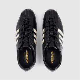 Adidas trainers in core black with cream white three stripes, gold branding detail, viewed from above, flat lay shot.