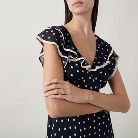 Model wearing Rixo Bethea Dot Dress in Layla Dot Navy, showing V-neckline with white-trimmed ruffles and polka dot pattern.