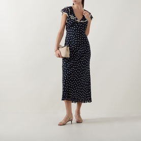 Model wearing Rixo Bethea Dot Dress in Layla Dot Navy, midi-length bias cut with ruffle V-neckline, styled with nude heels and cream clutch bag.