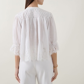 Model wearing Rixo ivory cotton top, rear view, with smocked yoke, elbow-length sleeves with lace trim, paired with white trousers.