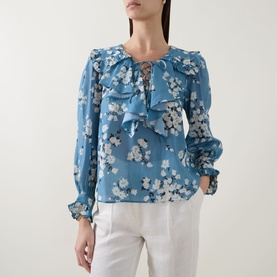 Model wearing Rixo top in Amelia Bouquet Blue floral print with ruffle neckline and lace-up detail, styled with white trousers.