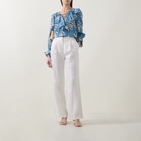 Model wearing Rixo Amelia Bouquet Blue floral top with ruffled neckline and long sleeves, styled with white wide-leg trousers and nude heels.