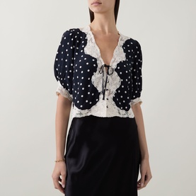 Model wearing Rixo navy polka dot viscose top with white lace trim, V-neck tie detail and puff sleeves, styled with a black satin skirt.