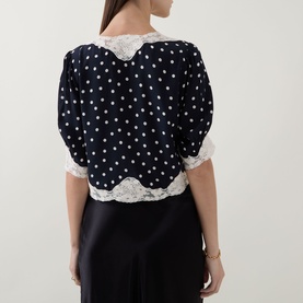 Model wearing Rixo navy polka dot viscose top, back view, with white lace collar and hem trim, styled with black satin skirt