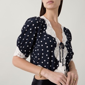 Model wearing Rixo top in Layla Dot Navy, white polka dot print with lace trim neckline and puff sleeves, gold bracelet visible.