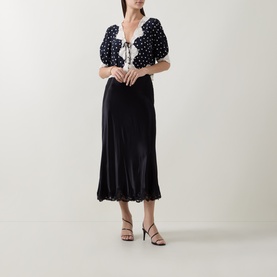 Model wearing Rixo Layla Dot Navy polka dot top with puff sleeves and lace trim, styled with a black midi skirt and strappy heels