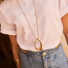 Model with gold-tone long chain necklace featuring an open oval loop pendant, styled with white tee and denim skirt