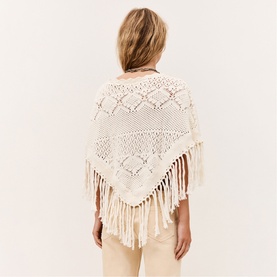 Model wearing Bash cream crochet knit poncho with diamond open-work pattern and long fringe hem, styled with cream trousers.