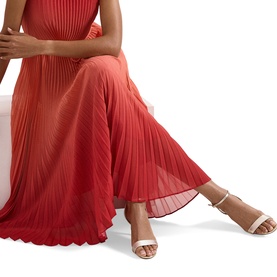 Model wearing Phase Eight red pleated maxi dress, styled with nude strappy heeled sandals, seated pose showing skirt detail.