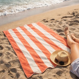 Allure Scallop Beach Towel in orange and white stripes laid on sandy beach, styled with a straw hat and sunglasses nearby