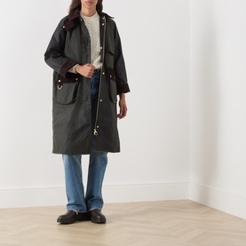 Model wearing Barbour Kirklees Wax Long Jacket in Fern, longline cut with corduroy collar and flap pockets, styled with blue jeans and black loafers