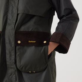 Model wearing Barbour Kirklees Wax Long Jacket in Fern, close-up of corduroy-trimmed flap pocket with gold logo buttons and burgundy cuff.