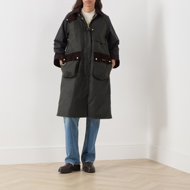 Model wearing Barbour Kirklees Wax Long Jacket in Fern, styled with blue jeans and black loafers. Longline cut with corduroy trim and flap pockets.